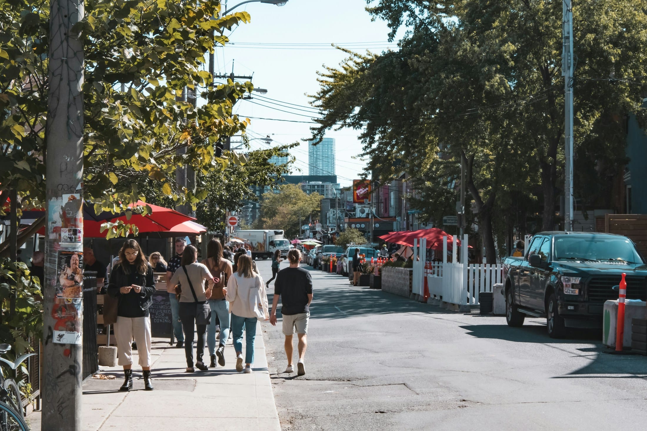 People walking along a sunny Kensington Market street in Toronto, past patio umbrellas and parked cars.